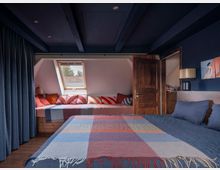 Dachgeschoss-Schlafzimmer mit dunkler blauer Decke und Wänden, einem großen Bett mit bunter Decke im Vordergrund und einer Fensternische im Hintergrund, ausgestattet mit Sitzpolstern und farbigen Kissen. Eine Holztür führt in einen angrenzenden Raum, während links ein blauer Vorhang zu erkennen ist.