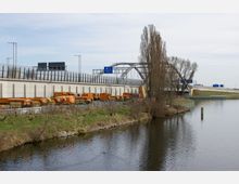 Eine moderne Straßenbrücke mit Lärmschutzwänden überspannt einen Fluss, daneben stehen zahlreiche orangefarbene Arbeitsfahrzeuge auf einem Parkplatz. Im Vordergrund ist eine Uferböschung mit Gras und Bäumen zu sehen.