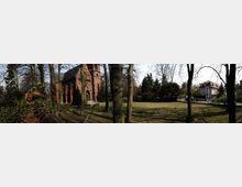 Panoramablick auf eine grüne Wiese mit vereinzelten Bäumen, im Hintergrund eine rote Backsteinkirche mit Spitzbogenfenstern und kleinem Turm sowie ein Wohnhaus mit hellem Putz und rotem Dach. Die Umgebung ist von Bäumen und Sträuchern umgeben, mit einem Zaun, der das Gelände abgrenzt.