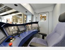 Innenansicht eines modernen Zugführerstands mit blauer Konsole, mehreren Bildschirmen, Schaltern und Steuerhebeln. Im Hintergrund befindet sich eine Tür mit einem kleinen Fenster, durch das Teile eines Lagerraums sichtbar sind.