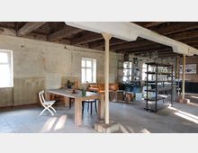 Ein rustikaler Innenraum mit unverputzten Wänden, freiliegenden Deckenbalken und Betonboden. Im Vordergrund stehen ein Holztisch mit verschiedenen Stühlen und Dekorationsgegenständen, im Hintergrund Sofas, ein Regal mit Gläsern und ein Tresenbereich.