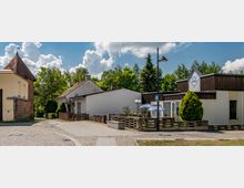 Straßenszene in einer kleinen Ortschaft mit Kopfsteinpflasterweg, einem gelben Gebäude mit Turmdach links und einem flachen, modernen Gebäude mit der Aufschrift „Li-Li Kino“ rechts. Im Hintergrund stehen Bäume unter einem bewölkten Himmel.