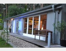 Flaches Gebäude mit grauer Holzfassade und großen Glasfronten, die einen Blick in den beleuchteten Innenraum ermöglichen. Vor dem Gebäude befinden sich ein Holzsteg, eine Holzbank, Kiesflächen und Pflanzen, im Hintergrund sind hohe Bäume sichtbar.