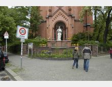 Backsteinkirche mit einer Statue Jesu vor dem Haupteingang, umgeben von grün bepflanzten Beeten und Blumen in Töpfen; im Vordergrund ein gepflasterter Gehweg, auf dem zwei Personen spazieren. Schilder weisen auf eine 30-km/h-Zone und Straßenschäden hin.