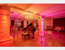 Eleganter Veranstaltungsraum mit Holzboden, weißen Stützsäulen und gewölbter Decke, in warmem Rot‑ und Violettlicht ausgeleuchtet. In der Mitte steht ein Flügel, umgeben von mehreren Kerzenleuchtern und einem Kronleuchter; im Hintergrund Fenster mit Vorhängen und eine Sitzecke.