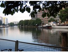 Stadtansicht mit einem ruhigen Fluss, an dessen Ufer Boote angelegt sind. Im Hintergrund befinden sich mehrstöckige Wohn- und Bürogebäude sowie Bäume, der Vordergrund wird von einem Zaun und überhängenden Baumblättern eingerahmt.