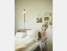 Ein minimalistisches Schlafzimmer mit einem Holzbett, weißen Bettlaken und einer gelben Decke. An der Wand hängen ein schlankes Wandlicht, zwei kleine dekorative Spiegel und gerahmte Fotografien; daneben steht eine silberne Statue auf einem transparenten Sockel.