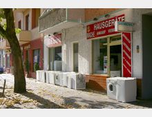 Straßenansicht eines Geschäfts für Haushaltsgeräte mit Schaufenstern und auffälligen roten Schildern. Vor dem Geschäft stehen gebrauchte Waschmaschinen und andere Geräte auf dem Gehweg; daneben ist ein Baum und der Boden ist mit Herbstlaub bedeckt.