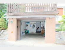 Garage mit offener Tür in einem Wohngebiet, mit brauner Putzfassade und der Hausnummer „15“. Im Inneren sind Wände aus weiß gestrichenem Mauerwerk, verschiedene Werkzeuge, Kabel, sowie eine Werkbank sichtbar.