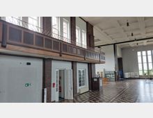 Großer Innenraum mit hohen Decken und großen Fenstern, Wänden aus Backstein und weißer Putzfläche. Ein Balkon mit Holzgeländer und Paneelen verläuft entlang der oberen Wand, der Boden ist mit einem geometrischen Fliesenmuster gestaltet.