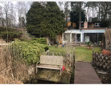 Ein Garten mit Rasenfläche, Holzzaun und Steg, der zu einer Holzbank führt. Im Hintergrund steht ein modernes Haus mit großen Fenstern, gelben Wänden und einem angebauten Wintergarten, umgeben von Bäumen und Sträuchern.