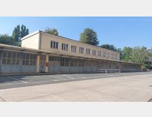 Langes, einstöckiges Gebäude mit beigem Putz und mehreren großen Fenstern in der oberen Etage; im unteren Bereich befinden sich zahlreiche graue Metalltüren, teils mit Treppen. Der Vorplatz ist asphaltiert, und im Hintergrund sind Bäume sichtbar.