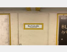 Ein U-Bahnschild mit der Aufschrift „Kochstraße Checkpoint Charlie“ an einer beige gestrichenen Wand, umrahmt von einem gelben Rahmen; daneben ein Plakatausschnitt mit Werbung.