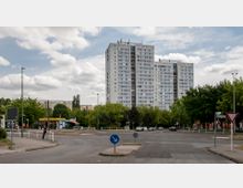Hochhaus mit der Aufschrift "Kienbergtower" in einem urbanen Wohngebiet, umgeben von Bäumen und kleineren Gebäuden. Im Vordergrund befinden sich eine Straßenkreuzung mit Verkehrszeichen und ein Kioskbereich.