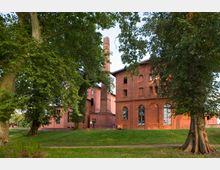 Backsteingebäude mit hohen Fenstern und einem Schornstein, umgeben von einer grünen Parklandschaft mit großen Bäumen und Rasenflächen. Im Vordergrund führen Wege durch die Grünfläche.