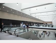 Großes, modernes Innenraumgebäude mit mehreren Ebenen, hell erleuchtet durch eine Reihe von Deckenleuchten und Tageslicht. Eine Rolltreppe verbindet die unteren Etagen mit einer Galerieebene, auf der Sitzbereiche und Personen zu sehen sind; glatt verputzte Wände und Glasgeländer prägen die Architektur.