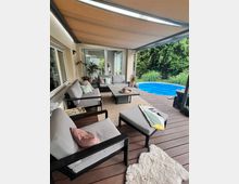 Überdachte Holzterrasse an einem Wohnhaus mit Lounge-Sitzmöbeln, Couchtisch und Teppich, angrenzend an einen runden, blauen Pool. Im Hintergrund stehen hohe, dichte Hecken und Ziergräser am Rand des Gartens.