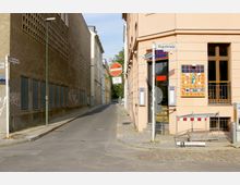 Straßenecke mit einer schmalen, asphaltierten Straße, flankiert von einem sandfarbenen Gebäude links mit vergitterten Fenstern und Graffiti, sowie einem hellbeigen Gebäude rechts mit farbig dekorierter Außenwand. Verkehrsschilder, darunter ein Durchfahrtverbots- und ein Fahrraderlaubnisschild, sind an der Straßenecke angebracht.