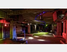 Industrieähnlicher Clubraum mit rohen Betonwänden, freiliegenden Rohren und niedriger Decke, beleuchtet von bunten Scheinwerfern und LED-Lichtstreifen in Blau. Der Raum hat erhöhte Plattformen, eine Tanzfläche in der Mitte und vereinzelte Lautsprecher an der Decke.