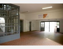 Ein Eingangsbereich mit rotem Fliesenboden, weißen gefliesten Wänden und einer Glastrennwand auf der linken Seite. Rechts befinden sich eine geöffnete Doppeltür und Werbeschilder über einer Durchgangsöffnung.