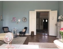 Wohnzimmer mit hellblauen Wänden, zwei schwarzen Sesseln, einem kleinen weißen Tisch und einer geöffneten zweiflügeligen Tür, die zu einem Flur mit Teppich führt. Der Raum ist mit Dekorationen wie Bildern, einem Spiegel und Blumen auf einem weißen Sideboard gestaltet. Im Vordergrund steht ein Tisch mit einem kleinen Standspiegel und Schmuck.