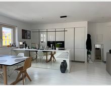 Moderner, lichtdurchfluteter Wohnraum mit offener Küche, weißen Hochglanzfronten und einer großen Kücheninsel. Im Vordergrund steht ein Esstisch mit Stühlen, während ein Fenster links bunte Fassaden eines Nachbargebäudes zeigt.