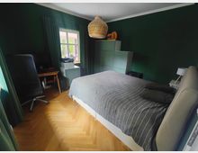 Schlafzimmer mit dunkelgrünen Wänden, Parkettboden und einem Doppelbett mit grauer Tagesdecke. Neben einem Fenster mit Vorhängen stehen ein Schreibtisch mit Computer und ein Drucker; gegenüber befindet sich ein hoher Schrank und eine geflochtene Pendelleuchte an der Decke.