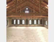 Innenraum eines historischen Gebäudes mit hohen, spitzbogigen Fenstern entlang einer ziegelsteinernen Wand. Der Raum verfügt über ein offenes Holzdach mit sichtbaren Balken und ein rundes Fenster im Giebel.