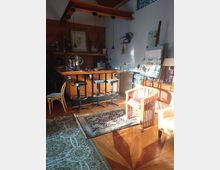 Innenraum eines gemütlichen Wohnraums mit einer hölzernen Bar, vier Barhockern und einem dekorativen Teppich auf einem Holzboden mit geometrischem Muster. Rechts befinden sich ein Holzstuhl mit rotem Kissen, ein Bücherstapel und Staffeleien, während links ein zweiter Teppich liegt.