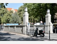 Straßenszene am U-Bahnhof Rüdesheimer Platz mit einem stilisierten Geländer aus Beton und zwei Laternenpfeilern, umgeben von Bäumen. Im Vordergrund ein geparkter Motorroller, im Hintergrund ein Wohngebäude mit Satteldach.