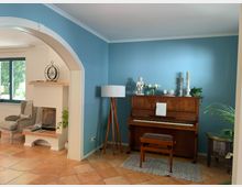 Wohnzimmer mit blauen Wänden und einem Klavier aus Holz, dekoriert mit weißen Kerzen und Blumenvasen. Im linken Bereich ein gemauerter Kamin, ein Sessel und ein großes Fenster, der Boden ist mit Terrakotta-Fliesen ausgelegt.