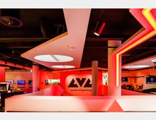 Gaming-Zentrum mit moderner Einrichtung, beleuchtet in Rot- und Orangetönen; im Hintergrund das Logo "LVL world of gaming" an einer Wand. Die Architektur umfasst weiße Säulen, offene Deckenkonstruktionen und geometrische Deckenplatten mit eingebauten Leuchten.