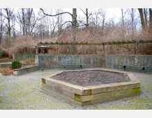 Öffentlicher Platz mit einer sechseckigen, hölzernen Hochbeet-Umrandung im Vordergrund, umgeben von gepflastertem Boden. Im Hintergrund befinden sich betonierte Sitzbänke, ein graffitibeschmierter Betonzaun und eine Holzpergola, daneben kahle Bäume und Sträucher in einer winterlichen Parklandschaft.