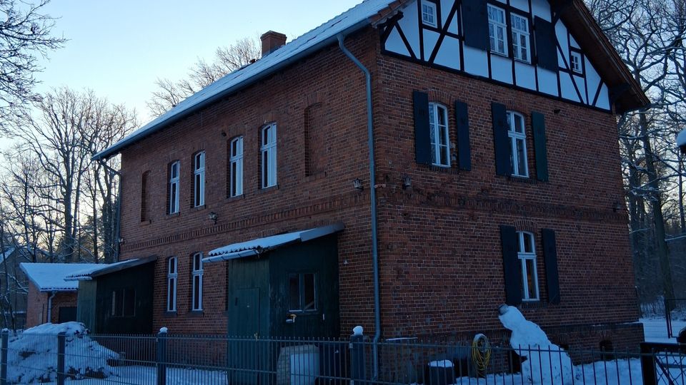 Backsteinbau eines zweigeschossigen Hauses mit Fachwerkelementen im oberen Giebelbereich, umgeben von einem verschneiten Gelände und einem Metallzaun. Im Hintergrund sind kahle Bäume und ein klarer Himmel zu sehen.