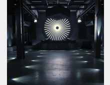 Ein dunkler, moderner Clubraum mit einer zentralen Bühne, die von einem großen, leuchtenden, radialen Lichtmuster hinterlegt wird. Der Boden ist aus glattem Beton, und die Beleuchtung erzeugt punktuelle Lichtreflexe auf dem Boden, während industrielle Elemente wie Säulen und sichtbare Deckenstrukturen den Raum definieren.