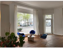 Heller Raum mit großen, bodentiefen Fenstern, weißen Vorhängen und einem Holzboden. Im Vordergrund stehen drei blaue Stühle um einen kleinen weißen Tisch, und Pflanzen sind links zu sehen; durch die Fenster ist eine Stadtstraße mit geparkten Autos zu erkennen.