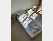 Helles Schlafzimmer mit einem Doppelbett aus Holz, das mit gestreifter Bettwäsche in Grau und Weiß bezogen ist. Auf beiden Seiten des Bettes befinden sich grüne Nachttischlampen, und durch ein Fenster fällt Sonnenlicht auf den beigefarbenen Teppichboden.