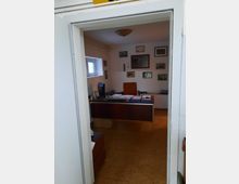 Blick in ein kleines Büro mit einem Holztisch, einem Computerbildschirm, einem orangefarbenen Bürostuhl und mehreren Dokumentenablagen. An den Wänden hängen gerahmte Bilder und Urkunden; Tageslicht fällt durch ein kleines Fenster ein.