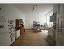 Geräumige Küche und Essbereich in einem hellen Raum mit Holzdielenboden. Links ein weißer Kühlschrank und ein Holzschrank, rechts eine Küchenzeile mit Oberschränken; im Hintergrund ein Esstisch mit blauer Tischdecke und Stühlen nahe einem großen Fenster mit Pflanzen.