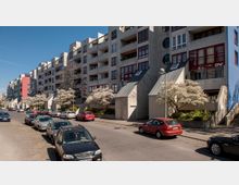 Wohnsiedlung mit modernen, mehrstöckigen Gebäuden aus Beton, roten Fensterrahmen und Balkonen. Entlang der Straße stehen geparkte Autos, und vor den Gebäuden blühen Bäume mit weißen Blüten.