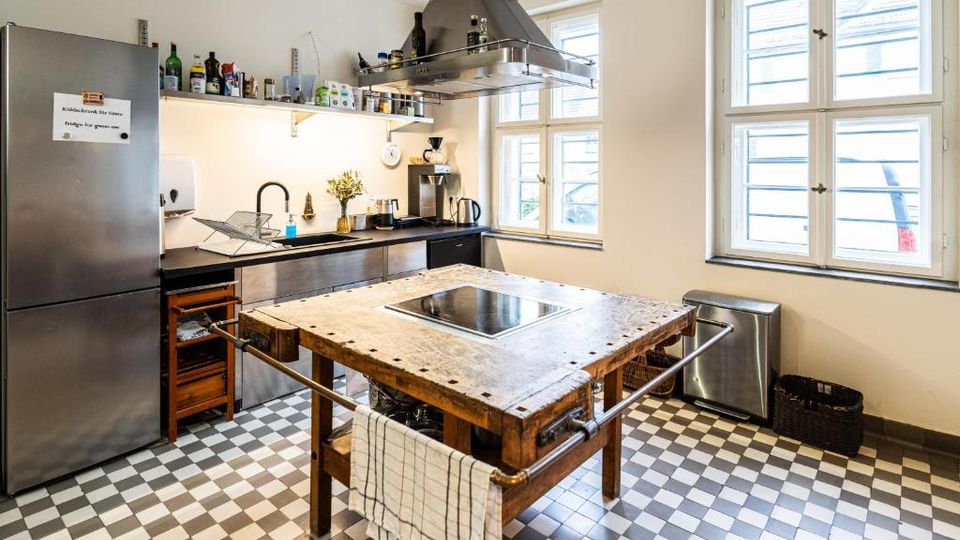 Helle Gemeinschaftsküche mit schwarz-weißen Schachbrettfliesen, Edelstahl-Kühlschrank und Küchenzeile mit Dunstabzugshaube. In der Mitte steht ein großer Holzarbeitstisch mit integriertem Kochfeld; große Fenster sorgen für viel Tageslicht.