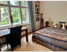 Helles Schlafzimmer mit großem Fenster, das auf einen grünen Außenbereich blickt. Der Raum ist eingerichtet mit einem Holztisch, einem schwarzen Stuhl, einem Bett mit gestreifter Decke, dekorativen Kissen, einer Bücherwand und einer Tischlampe auf einem Nachttisch.