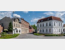 Straßenszene in einer kleinen Stadt mit historischen Gebäuden; links ein älteres, dreistöckiges Haus mit verblassender Schrift und Zierfassade, rechts ein weißes Gebäude mit rotem Ziegeldach. Im Vordergrund führt eine leicht geschwungene Straße durch einen begrünten Bereich.