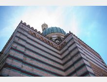 Eckansicht einer historischen Moschee mit gestreiftem Mauerwerk in rot-weißen und bläulichen Farbtönen, einem grünlichen Kuppeldach und einem kleinen Minarett im Hintergrund unter klarem Himmel.