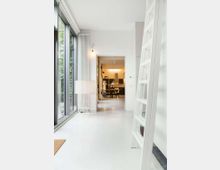 Bright modern hallway with white floors and walls, a tall window wall on the left, and a leaning white ladder on the right. The corridor leads through an open doorway to a warmly lit kitchen and dining area with pendant lights and dark cabinetry.