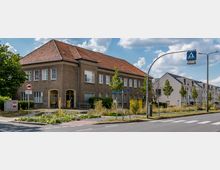 Straßenszene mit einem zweistöckigen Gebäude im historischen Stil, roten Ziegeldach und mehreren weißen Fenstern. Im Vordergrund befinden sich eine Straßenkreuzung mit Zebrastreifen, Verkehrszeichen und eine kleine Grünfläche mit Bäumen und Blumen.