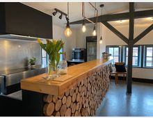 Moderne Küche mit einer Theke aus Holzscheiten, darauf eine Vase mit Tulpen und ein Glas. Im Hintergrund sind große Fenster, schwarze Stahlträger, Hängeleuchten und eine Edelstahlküche zu sehen.