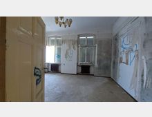 Ein leerer, heruntergekommener Raum mit hohen Fenstern, beschädigten Wänden und Graffiti. Die Wände sind teilweise mit abblätterndem Putz bedeckt, und an der Decke hängt ein Kronleuchter mit mehreren Glühbirnen.