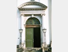 Eingangsbereich eines historischen Gebäudes mit einer grünen Holztür, einem halbrunden Fenster darüber und einem verzierten Steinportal mit Säulen und Gesimsen. Zwei Laternen stehen jeweils links und rechts der Steintreppe.