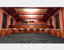 Historischer Theatersaal mit roten Wänden und Holzelementen, mehreren Sitzreihen, einer Empore und einer flachen Decke mit rechteckigen Lampen. Große Vorhänge bedecken Fenster an beiden Seitenwänden.
